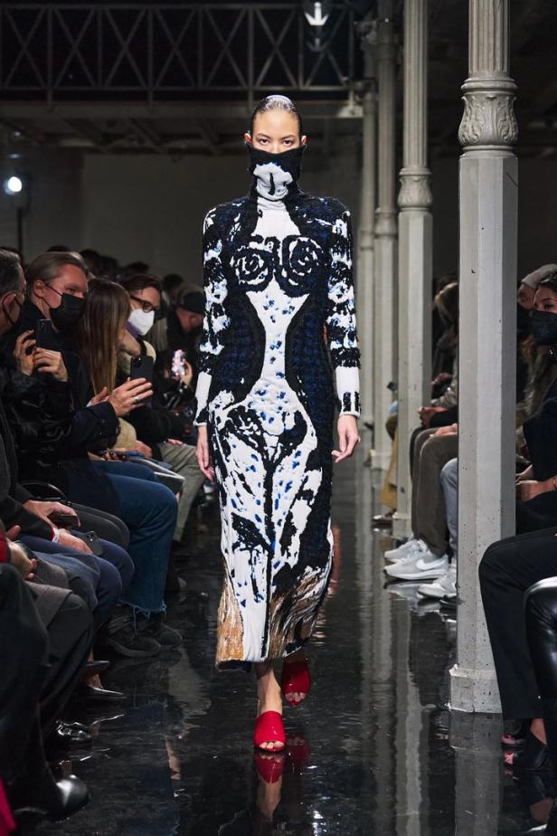 Ein Model präsentiert ein schwarz-weiß-blaues Kleid mit Rollkragen und Gesichtsbedeckung auf dem Laufsteg.