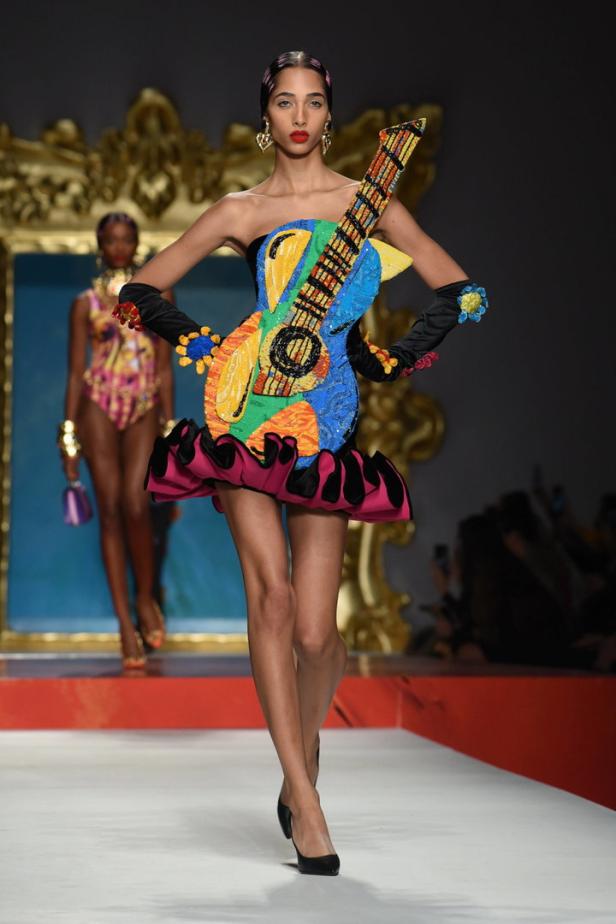 Ein Model präsentiert ein farbenfrohes Kleid in Gitarrenform auf einem Laufsteg.