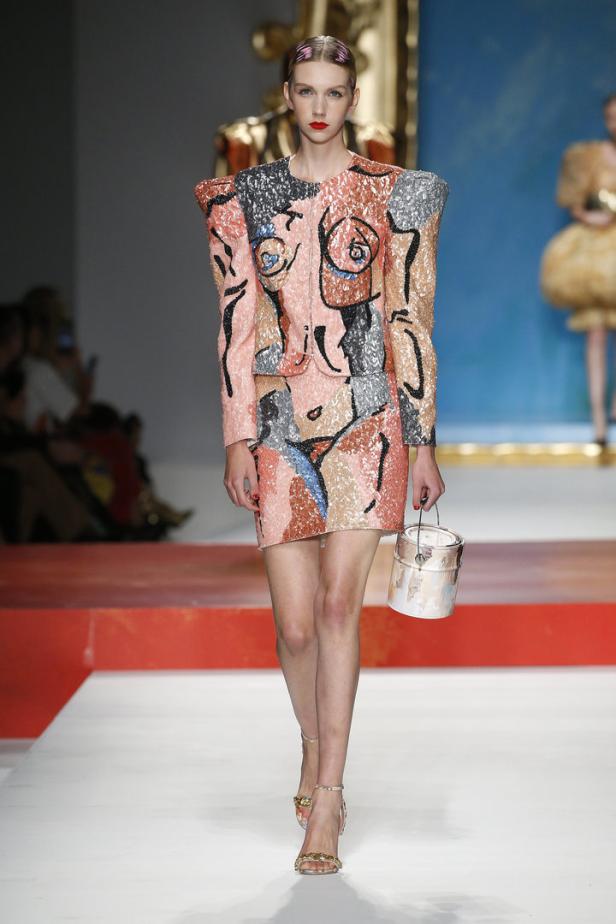 Ein Model auf dem Laufsteg in einem Kleid mit abstrakten Körperzeichnungen und einer Farbdosen-Handtasche.