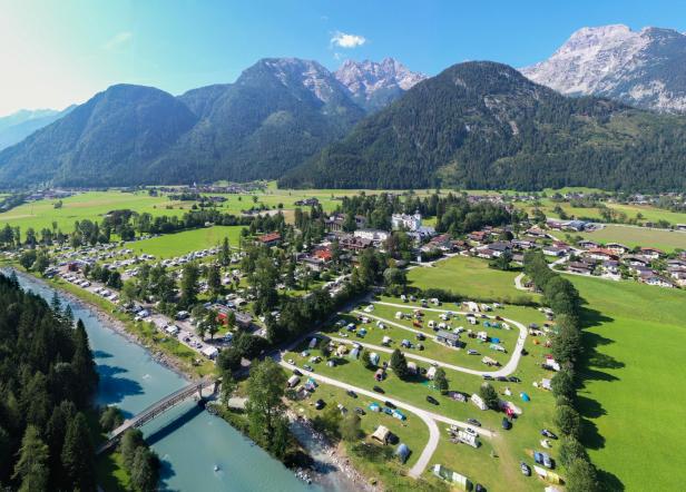 Luftaufnahme eines Campingplatzes am Flussufer mit Bergen im Hintergrund.