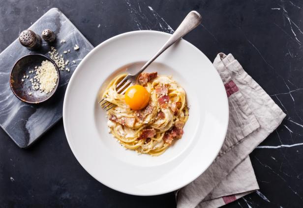 Ein Teller Pasta Carbonara mit einem Eigelb und Speckwürfeln wird mit einer Gabel angerichtet.