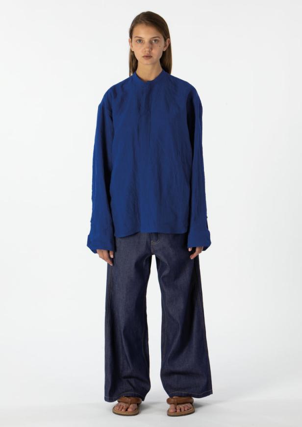 Eine Frau trägt eine blaue Bluse und weite Jeans vor einem weißen Hintergrund.