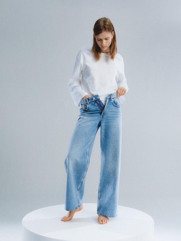 Eine Frau mit einem weißen Oberteil und einer offenen, hellblauen Jeans steht barfuß auf einem Podest.