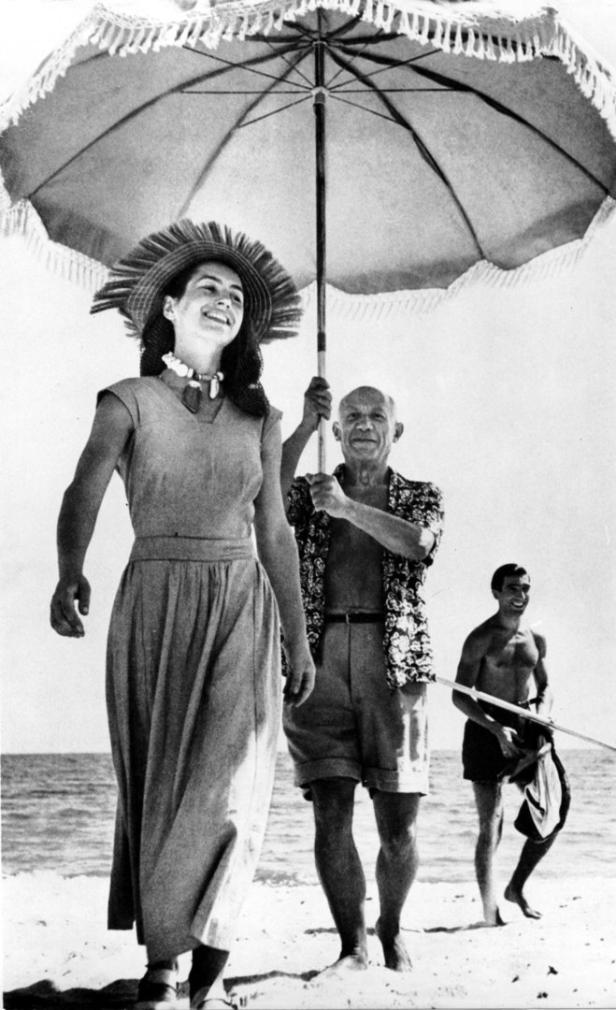 Pablo Picasso hält einen Sonnenschirm über einer Frau am Strand, während ein Mann im Hintergrund steht.
