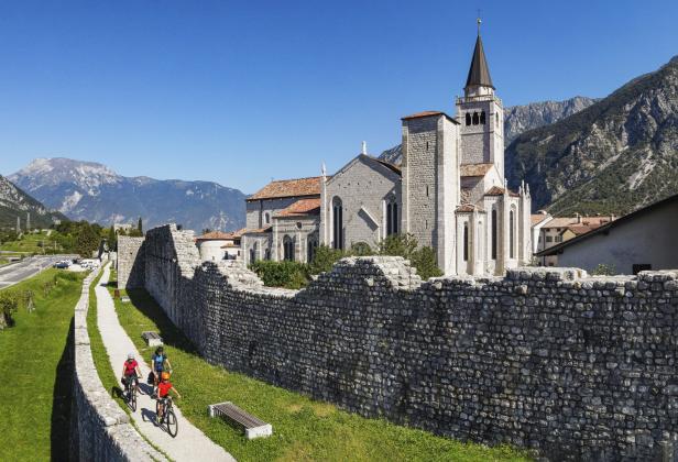 Drei Radfahrer fahren auf einem Weg entlang einer alten Steinmauer mit einer Kirche im Hintergrund.