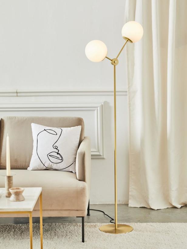 Eine goldene Stehlampe mit zwei kugelförmigen Lampenschirmen neben einem Sofa mit Kissen.