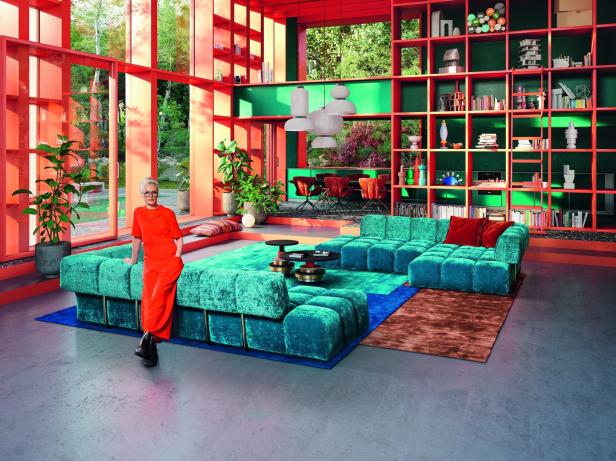 Eine Frau in einem orangefarbenen Overall lehnt an einem türkisfarbenen Sofa in einem farbenfrohen Wohnzimmer.