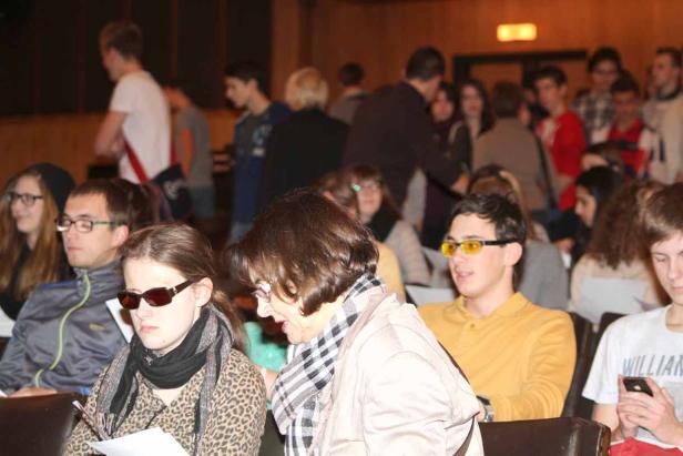 Zuschauer sitzen in einem Raum, einige tragen Sonnenbrillen.