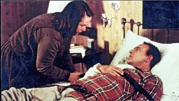 Eine Frau beugt sich über einen Mann, der im Bett liegt und eine Verletzung hat.