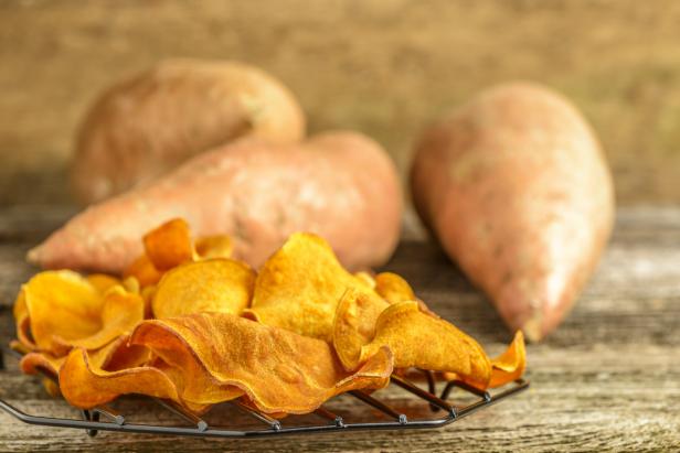 Süßkartoffelchips liegen vor rohen Süßkartoffeln auf einem Holzbrett.