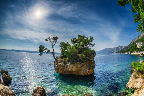 Ein kleiner Felsen mit Bäumen im türkisfarbenen Wasser vor einer Küstenlandschaft.