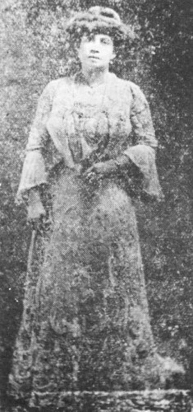 Eine Frau in einem verzierten Kleid und Hut steht vor einem dunklen Hintergrund.