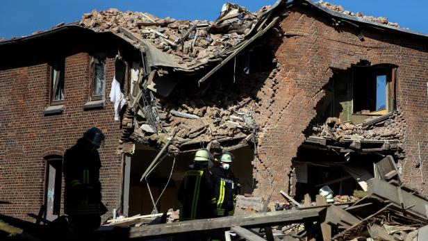 Ein beschädigtes Backsteingebäude mit Trümmern und Feuerwehrleuten vor Ort.
