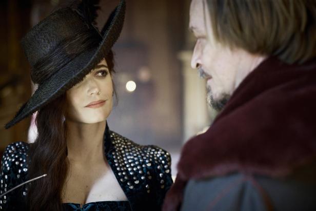 Eva Green in einem dunklen Kleid und Hut blickt einen Mann mit Bart an.
