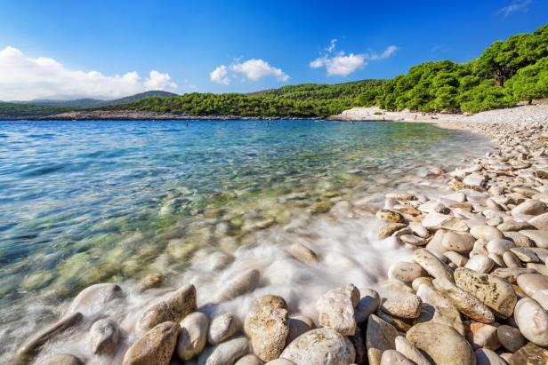 Ein Strand mit klarem Wasser und einem bewaldeten Hügel im Hintergrund.