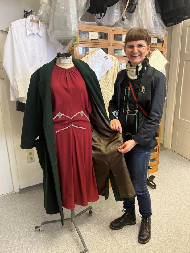 Eine Frau steht neben einer Schneiderpuppe mit einem roten Kleid und einem grünen Mantel.
