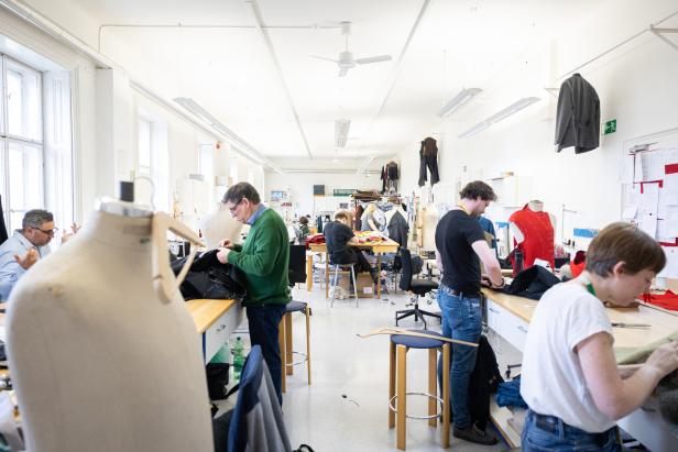 In einem hellen Atelier arbeiten mehrere Personen an Tischen mit Stoffen und Schneiderpuppen.