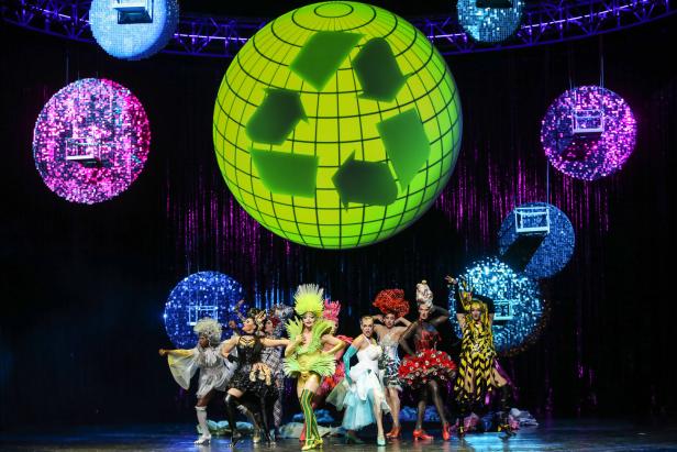 Eine Gruppe von Drag Queens steht auf einer Bühne vor einem Globus mit einem Recycling-Symbol.