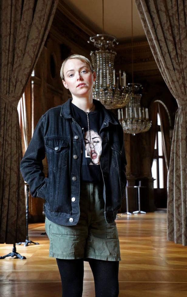 Eine junge Frau mit kurzer, blonder Haaren, gekleidet in Jeansjacke und Shorts, steht in einem eleganten Raum.