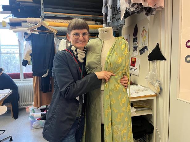 Eine Schneiderin posiert mit einer Schneiderpuppe, die ein grünes Kleid mit goldenem Muster trägt.