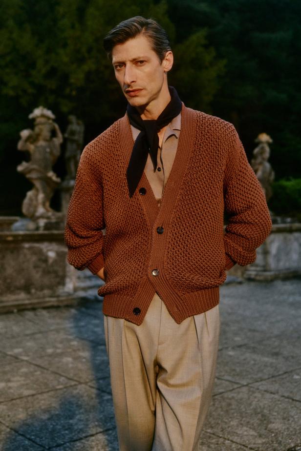 Ein Mann mit brauner Strickjacke und Halstuch posiert im Freien.
