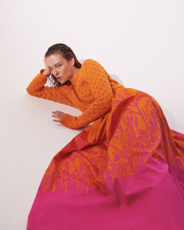 Eine Frau liegt in einem orangefarbenen Pullover und einem pinkfarbenen Rock mit floralem Muster auf dem Boden.