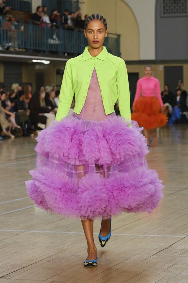 Eine Frau in einem pinkfarbenen Tüllrock und einer hellgrünen Jacke auf einem Laufsteg.