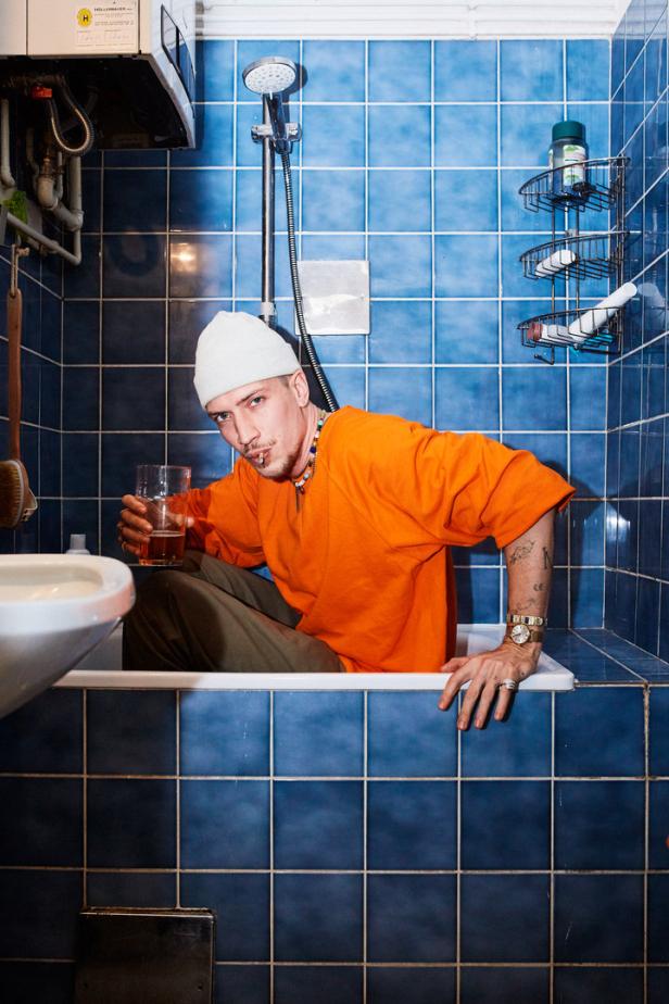 Ein Mann mit Mütze sitzt in einer blau gefliesten Badewanne und hält ein Glas Bier.