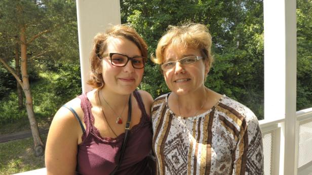 Zwei Frauen stehen lächelnd auf einem Balkon vor Bäumen.