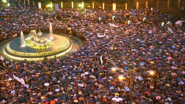 Eine große Menschenmenge mit Regenschirmen versammelt sich um einen beleuchteten Brunnen.