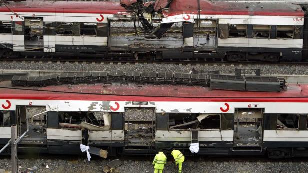 Zwei beschädigte rote und weiße Züge stehen auf den Gleisen, während zwei Arbeiter in gelben Anzügen die Trümmer untersuchen.