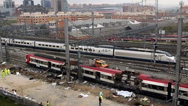 Ein zerstörter Zug steht auf den Gleisen, während im Hintergrund weitere Züge und Gebäude zu sehen sind.