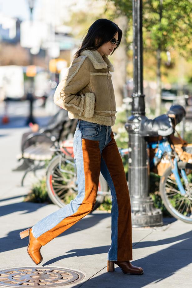 Eine Frau mit Sonnenbrille, Shearling-Jacke, Jeans mit braunen Wildledereinsätzen und braunen Stiefeln geht die Straße entlang.