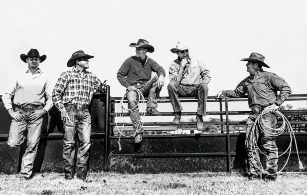 Fünf Cowboys sitzen oder stehen an einem Zaun, einige mit Lassos.