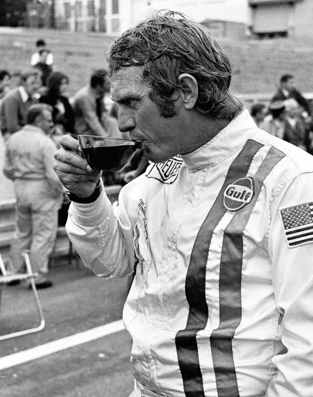 Steve McQueen trinkt in seiner Rennfahreruniform einen Schluck aus einer Tasse.
