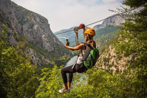 Eine Frau fährt mit einer Seilrutsche über eine grüne Schlucht und macht ein Selfie.