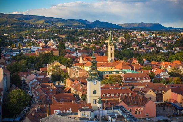 Blick über Zagreb mit roten Dächern und Kirchtürmen vor bewaldeten Hügeln.