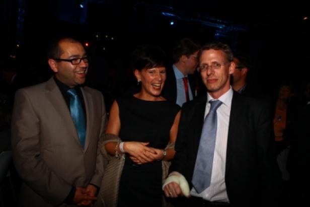 Drei Personen, gekleidet in Anzügen und einem Kleid, lachen zusammen.