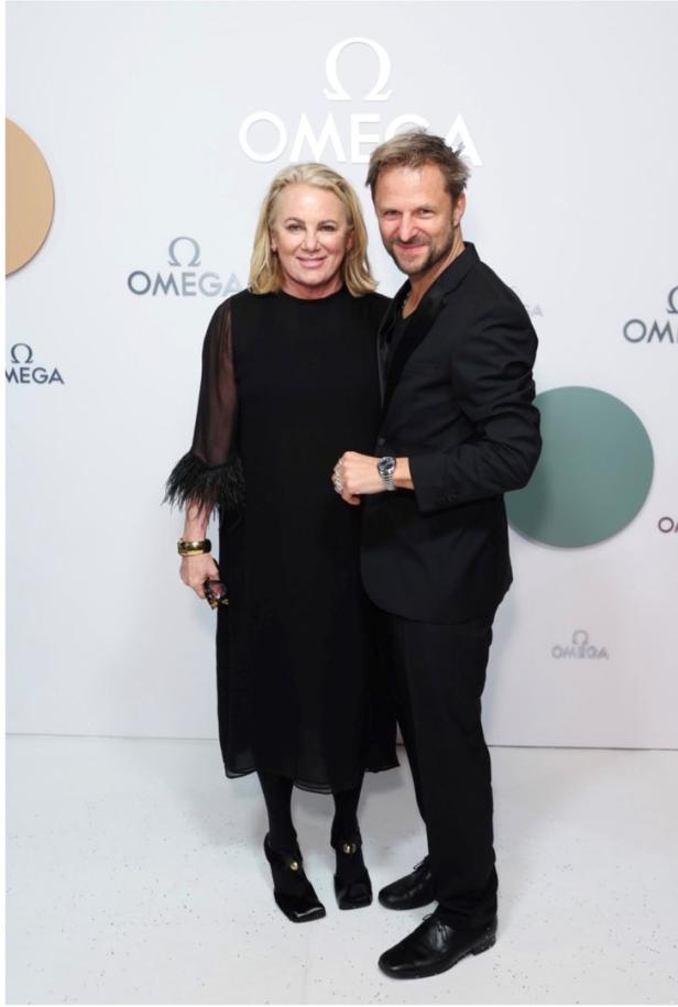 Zwei Personen posieren vor einem Omega-Logo an einer Wand.