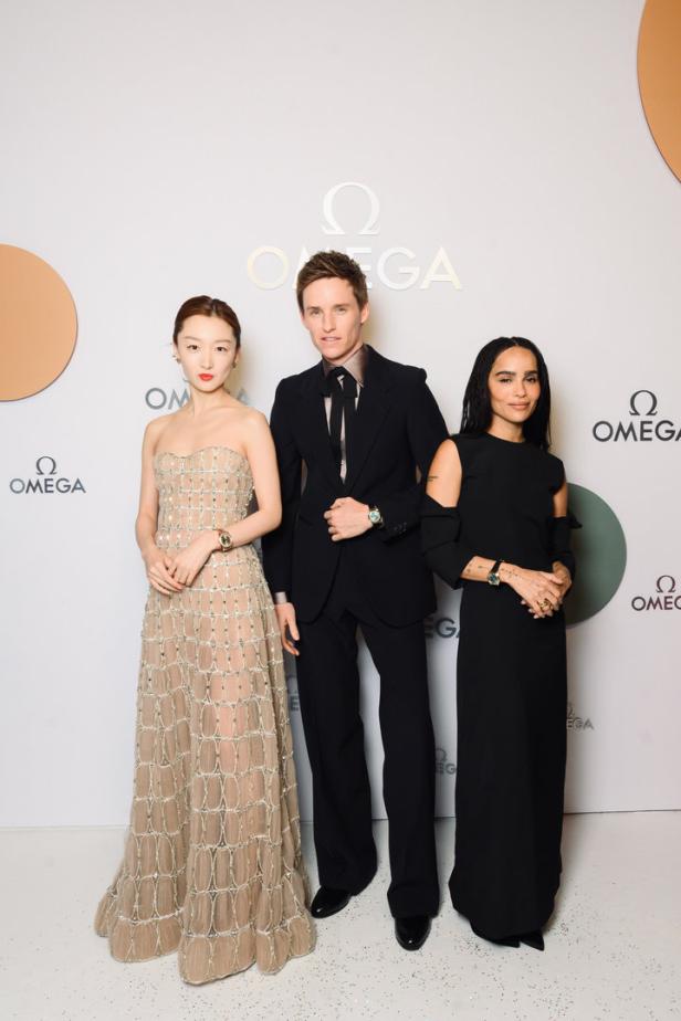 Drei Personen posieren vor einem Omega-Logo: eine Frau in einem beigen Kleid, Eddie Redmayne und Zoë Kravitz in einem schwarzen Kleid.