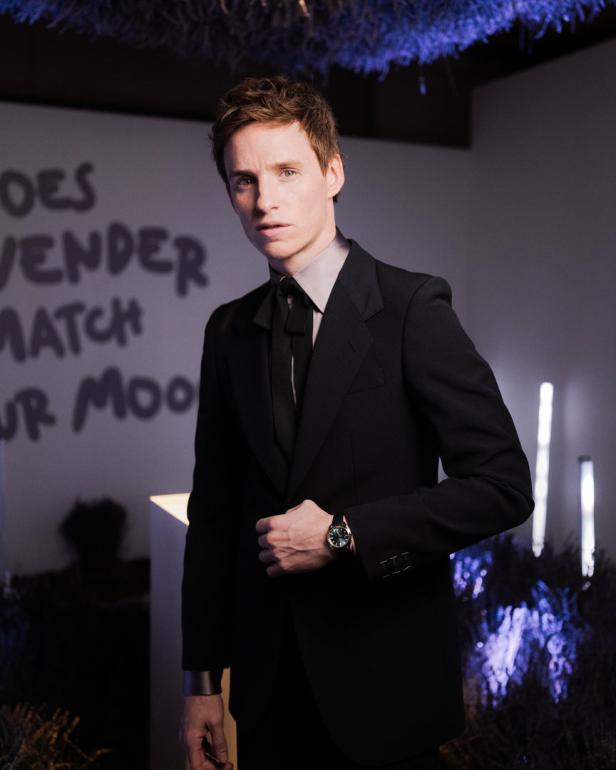 Eddie Redmayne in einem schwarzen Anzug mit einer Uhr am Handgelenk.