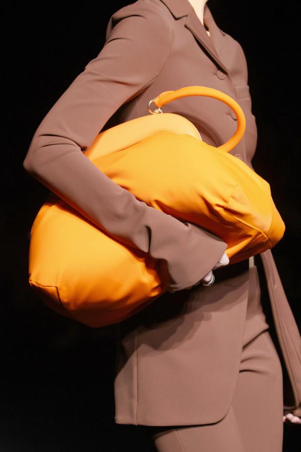 Ein Model trägt eine übergroße, orangefarbene Handtasche und einen braunen Hosenanzug.