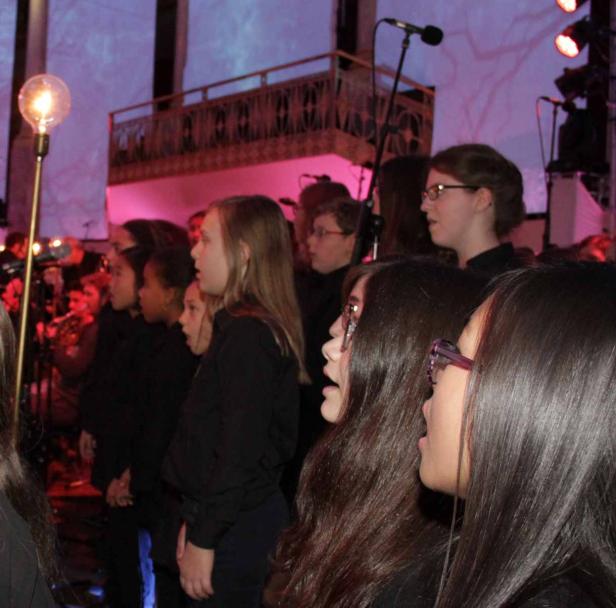 Ein Mädchenchor singt auf einer Bühne mit roter Beleuchtung.