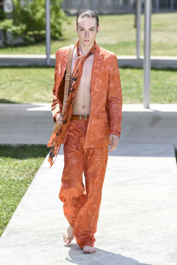 Ein barfuß laufendes Model präsentiert einen orangefarbenen Anzug mit Blumenmuster.