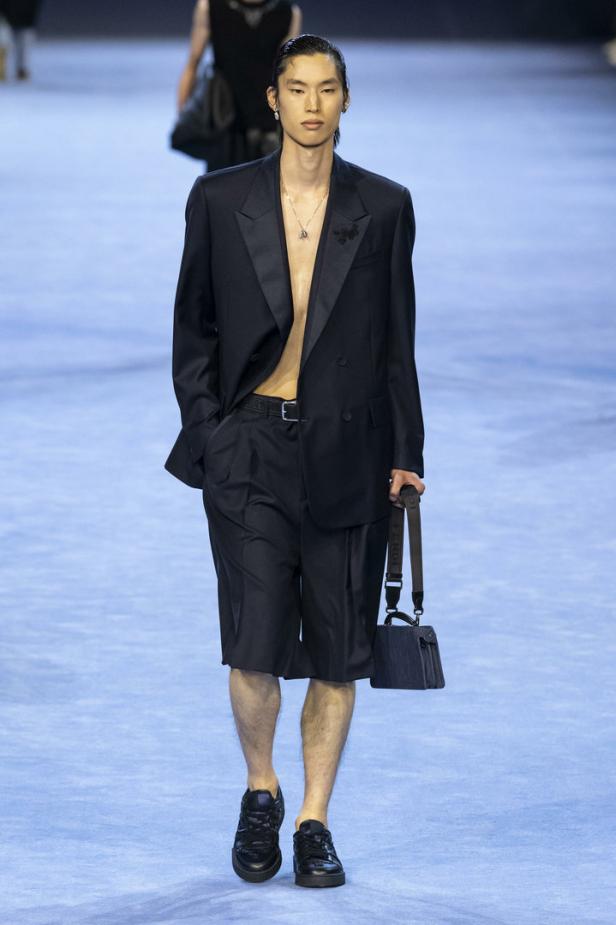 Ein Mann in einem offenen Blazer, Shorts und Turnschuhen auf einem Laufsteg mit einer kleinen Handtasche.