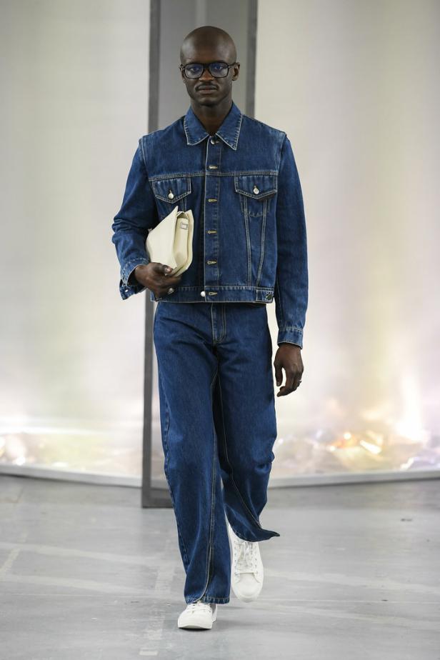 Ein Mann mit Brille trägt ein Jeans-Outfit und eine helle Handtasche.