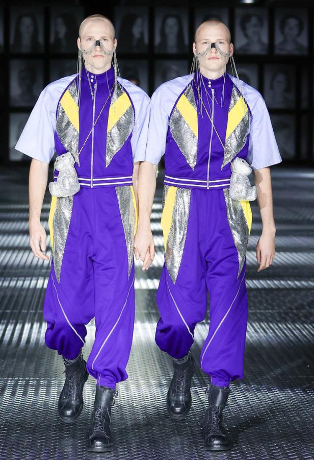 Zwei Models in lila Trainingsanzügen mit silbernen Applikationen und Teddybär-Taschen auf einem Laufsteg.