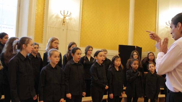 Ein Chor von Mädchen probt unter der Leitung eines Dirigenten in einem Raum mit goldener Tapete.