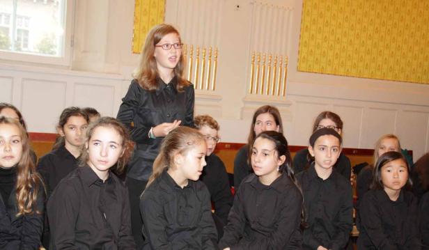 Ein Mädchen leitet einen Kinderchor in einem Raum mit gelber Tapete.