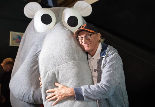 Ein Mann mit Brille und Baseballkappe umarmt eine große, graue Nilpferd-Figur.